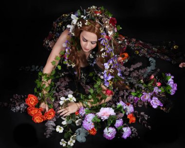 Celtic Bahar Tanrıçası Olarak Kadın Model Stüdyo Portreleri, Beltane Pagan Mitolojisinden esinlenerek Çiçek Fantezi Pelerini Kostümü. Klasik oturma pozu çiçek yatağında, izole edilmiş Karanlık Arkaplan