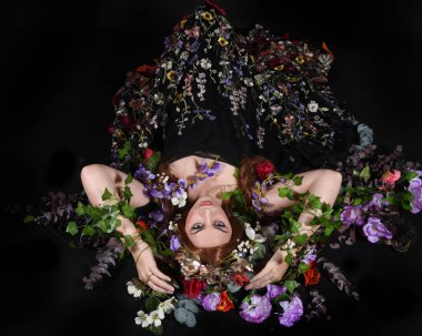 Celtic Bahar Tanrıçası Olarak Kadın Model Stüdyo Portreleri, Beltane Pagan Mitolojisinden esinlenerek Çiçek Fantezi Pelerini Kostümü. Klasik oturma pozu çiçek yatağında, izole edilmiş Karanlık Arkaplan