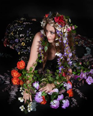 Celtic Bahar Tanrıçası Olarak Kadın Model Stüdyo Portreleri, Beltane Pagan Mitolojisinden esinlenerek Çiçek Fantezi Pelerini Kostümü. Klasik oturma pozu çiçek yatağında, izole edilmiş Karanlık Arkaplan