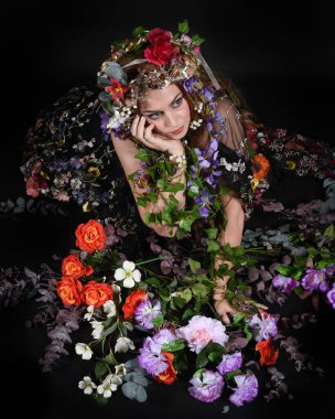 Celtic Bahar Tanrıçası Olarak Kadın Model Stüdyo Portreleri, Beltane Pagan Mitolojisinden esinlenerek Çiçek Fantezi Pelerini Kostümü. Klasik oturma pozu çiçek yatağında, izole edilmiş Karanlık Arkaplan