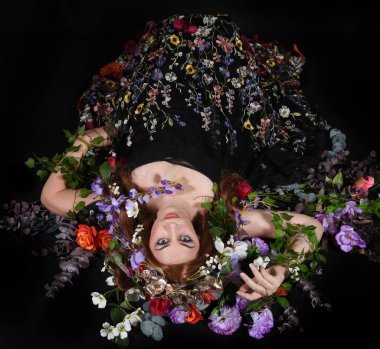 Celtic Bahar Tanrıçası Olarak Kadın Model Stüdyo Portreleri, Beltane Pagan Mitolojisinden esinlenerek Çiçek Fantezi Pelerini Kostümü. Klasik oturma pozu çiçek yatağında, izole edilmiş Karanlık Arkaplan