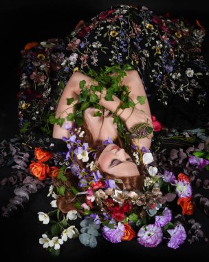 Celtic Bahar Tanrıçası Olarak Kadın Model Stüdyo Portreleri, Beltane Pagan Mitolojisinden esinlenerek Çiçek Fantezi Pelerini Kostümü. Klasik oturma pozu çiçek yatağında, izole edilmiş Karanlık Arkaplan