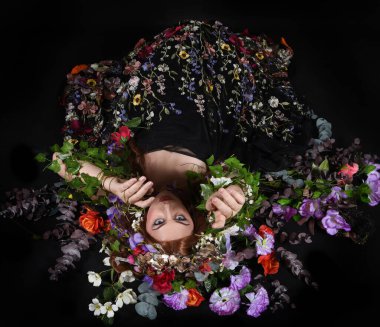 Celtic Bahar Tanrıçası Olarak Kadın Model Stüdyo Portreleri, Beltane Pagan Mitolojisinden esinlenerek Çiçek Fantezi Pelerini Kostümü. Klasik oturma pozu çiçek yatağında, izole edilmiş Karanlık Arkaplan