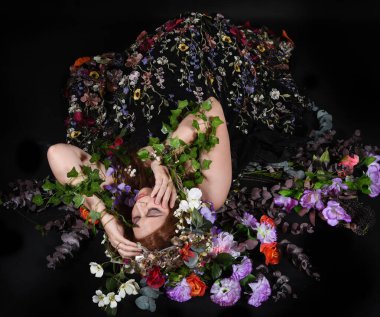 Celtic Bahar Tanrıçası Olarak Kadın Model Stüdyo Portreleri, Beltane Pagan Mitolojisinden esinlenerek Çiçek Fantezi Pelerini Kostümü. Klasik oturma pozu çiçek yatağında, izole edilmiş Karanlık Arkaplan