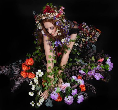 Celtic Bahar Tanrıçası Olarak Kadın Model Stüdyo Portreleri, Beltane Pagan Mitolojisinden esinlenerek Çiçek Fantezi Pelerini Kostümü. Klasik oturma pozu çiçek yatağında, izole edilmiş Karanlık Arkaplan