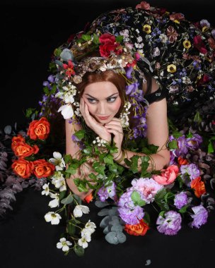 Celtic Bahar Tanrıçası Olarak Kadın Model Stüdyo Portreleri, Beltane Pagan Mitolojisinden esinlenerek Çiçek Fantezi Pelerini Kostümü. Klasik oturma pozu çiçek yatağında, izole edilmiş Karanlık Arkaplan