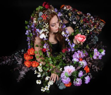 Celtic Bahar Tanrıçası Olarak Kadın Model Stüdyo Portreleri, Beltane Pagan Mitolojisinden esinlenerek Çiçek Fantezi Pelerini Kostümü. Klasik oturma pozu çiçek yatağında, izole edilmiş Karanlık Arkaplan