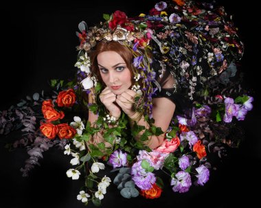 Celtic Bahar Tanrıçası Olarak Kadın Model Stüdyo Portreleri, Beltane Pagan Mitolojisinden esinlenerek Çiçek Fantezi Pelerini Kostümü. Klasik oturma pozu çiçek yatağında, izole edilmiş Karanlık Arkaplan