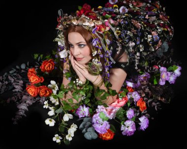 Celtic Bahar Tanrıçası Olarak Kadın Model Stüdyo Portreleri, Beltane Pagan Mitolojisinden esinlenerek Çiçek Fantezi Pelerini Kostümü. Klasik oturma pozu çiçek yatağında, izole edilmiş Karanlık Arkaplan