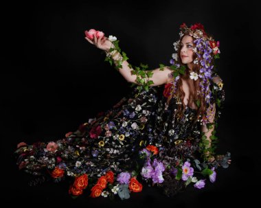 Celtic Bahar Tanrıçası Olarak Kadın Model Stüdyo Portreleri, Beltane Pagan Mitolojisinden esinlenerek Çiçek Fantezi Pelerini Kostümü. Klasik oturma pozisyonu, kollar uzanıyor. izole edilmiş Karanlık Arkaplan