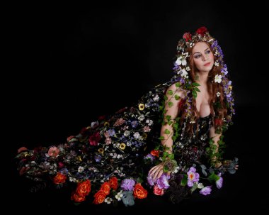 Celtic Bahar Tanrıçası Olarak Kadın Model Stüdyo Portreleri, Beltane Pagan Mitolojisinden esinlenerek Çiçek Fantezi Pelerini Kostümü. Klasik oturma pozisyonu, kollar uzanıyor. izole edilmiş Karanlık Arkaplan