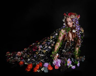 Celtic Bahar Tanrıçası Olarak Kadın Model Stüdyo Portreleri, Beltane Pagan Mitolojisinden esinlenerek Çiçek Fantezi Pelerini Kostümü. Klasik oturma pozisyonu, kollar uzanıyor. izole edilmiş Karanlık Arkaplan