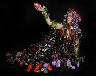 Celtic Bahar Tanrıçası Olarak Kadın Model Stüdyo Portreleri, Beltane Pagan Mitolojisinden esinlenerek Çiçek Fantezi Pelerini Kostümü. Klasik oturma pozisyonu, kollar uzanıyor. izole edilmiş Karanlık Arkaplan