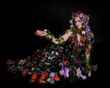 Celtic Bahar Tanrıçası Olarak Kadın Model Stüdyo Portreleri, Beltane Pagan Mitolojisinden esinlenerek Çiçek Fantezi Pelerini Kostümü. Klasik oturma pozisyonu, kollar uzanıyor. izole edilmiş Karanlık Arkaplan