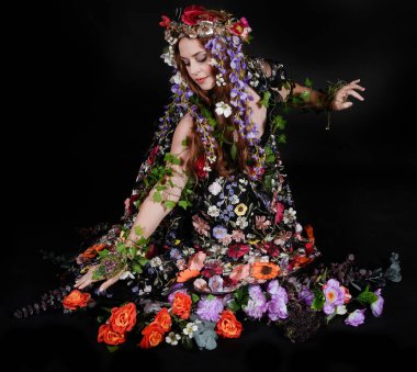 Celtic Bahar Tanrıçası Olarak Kadın Model Stüdyo Portreleri, Beltane Pagan Mitolojisinden esinlenerek Çiçek Fantezi Pelerini Kostümü. Klasik oturma pozisyonu, kollar uzanıyor. izole edilmiş Karanlık Arkaplan