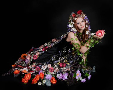 Celtic Bahar Tanrıçası Olarak Kadın Model Stüdyo Portreleri, Beltane Pagan Mitolojisinden esinlenerek Çiçek Fantezi Pelerini Kostümü. Klasik oturma pozisyonu, kollar uzanıyor. izole edilmiş Karanlık Arkaplan