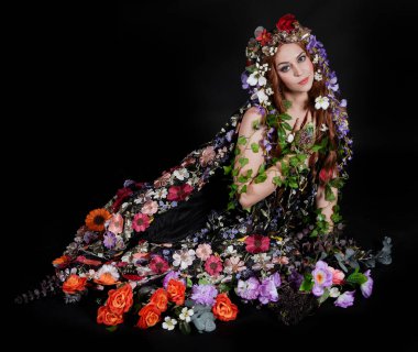 Celtic Bahar Tanrıçası Olarak Kadın Model Stüdyo Portreleri, Beltane Pagan Mitolojisinden esinlenerek Çiçek Fantezi Pelerini Kostümü. Klasik oturma pozisyonu, kollar uzanıyor. izole edilmiş Karanlık Arkaplan