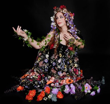 Celtic Bahar Tanrıçası Olarak Kadın Model Stüdyo Portreleri, Beltane Pagan Mitolojisinden esinlenerek Çiçek Fantezi Pelerini Kostümü. Klasik oturma pozisyonu, kollar uzanıyor. izole edilmiş Karanlık Arkaplan