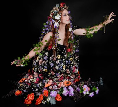 Celtic Bahar Tanrıçası Olarak Kadın Model Stüdyo Portreleri, Beltane Pagan Mitolojisinden esinlenerek Çiçek Fantezi Pelerini Kostümü. Klasik oturma pozisyonu, kollar uzanıyor. izole edilmiş Karanlık Arkaplan