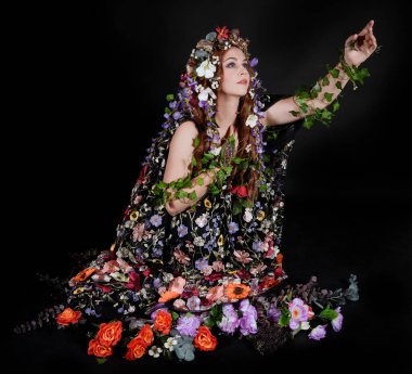 Celtic Bahar Tanrıçası Olarak Kadın Model Stüdyo Portreleri, Beltane Pagan Mitolojisinden esinlenerek Çiçek Fantezi Pelerini Kostümü. Klasik oturma pozisyonu, kollar uzanıyor. izole edilmiş Karanlık Arkaplan