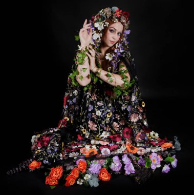 Celtic Bahar Tanrıçası Olarak Kadın Model Stüdyo Portreleri, Beltane Pagan Mitolojisinden esinlenerek Çiçek Fantezi Pelerini Kostümü. Klasik oturma pozisyonu, kollar uzanıyor. izole edilmiş Karanlık Arkaplan