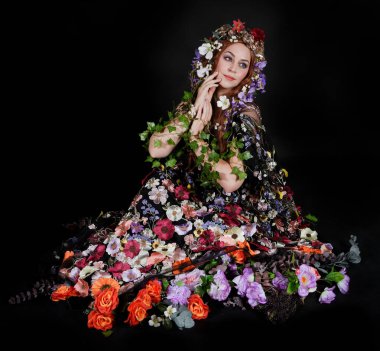 Celtic Bahar Tanrıçası Olarak Kadın Model Stüdyo Portreleri, Beltane Pagan Mitolojisinden esinlenerek Çiçek Fantezi Pelerini Kostümü. Klasik oturma pozisyonu, kollar uzanıyor. izole edilmiş Karanlık Arkaplan