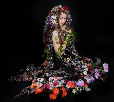 Celtic Bahar Tanrıçası Olarak Kadın Model Stüdyo Portreleri, Beltane Pagan Mitolojisinden esinlenerek Çiçek Fantezi Pelerini Kostümü. Klasik oturma pozisyonu, kollar uzanıyor. izole edilmiş Karanlık Arkaplan