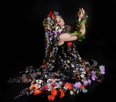 Celtic Bahar Tanrıçası Olarak Kadın Model Stüdyo Portreleri, Beltane Pagan Mitolojisinden esinlenerek Çiçek Fantezi Pelerini Kostümü. Klasik oturma pozisyonu, kollar uzanıyor. izole edilmiş Karanlık Arkaplan