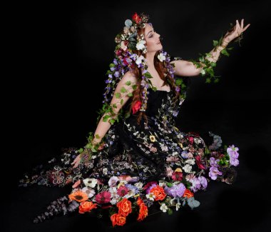 Celtic Bahar Tanrıçası Olarak Kadın Model Stüdyo Portreleri, Beltane Pagan Mitolojisinden esinlenerek Çiçek Fantezi Pelerini Kostümü. Klasik oturma pozisyonu, kollar uzanıyor. izole edilmiş Karanlık Arkaplan