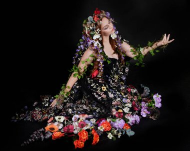 Celtic Bahar Tanrıçası Olarak Kadın Model Stüdyo Portreleri, Beltane Pagan Mitolojisinden esinlenerek Çiçek Fantezi Pelerini Kostümü. Klasik oturma pozisyonu, kollar uzanıyor. izole edilmiş Karanlık Arkaplan