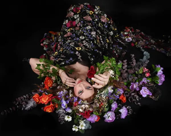 Celtic Bahar Tanrıçası Olarak Kadın Model Stüdyo Portreleri, Beltane Pagan Mitolojisinden esinlenerek Çiçek Fantezi Pelerini Kostümü. Klasik oturma pozu çiçek yatağında, izole edilmiş Karanlık Arkaplan