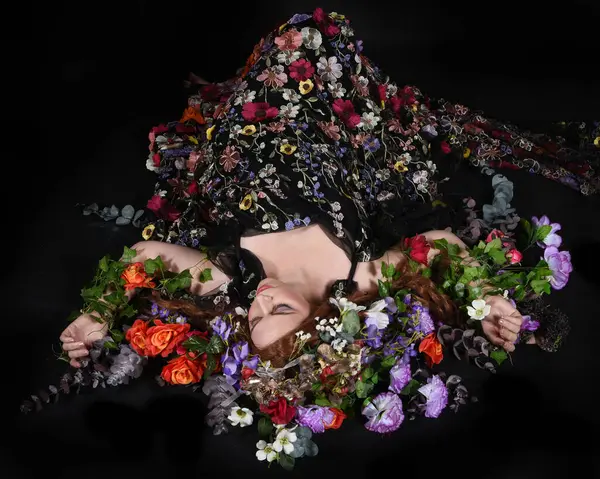 Celtic Bahar Tanrıçası Olarak Kadın Model Stüdyo Portreleri, Beltane Pagan Mitolojisinden esinlenerek Çiçek Fantezi Pelerini Kostümü. Klasik oturma pozu çiçek yatağında, izole edilmiş Karanlık Arkaplan