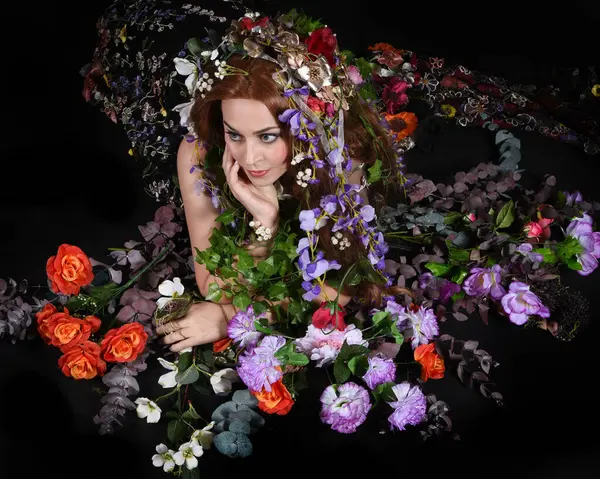 Celtic Bahar Tanrıçası Olarak Kadın Model Stüdyo Portreleri, Beltane Pagan Mitolojisinden esinlenerek Çiçek Fantezi Pelerini Kostümü. Klasik oturma pozu çiçek yatağında, izole edilmiş Karanlık Arkaplan