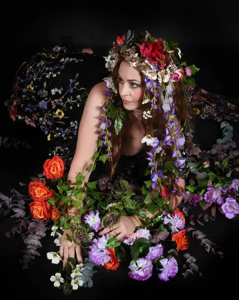 Celtic Bahar Tanrıçası Olarak Kadın Model Stüdyo Portreleri, Beltane Pagan Mitolojisinden esinlenerek Çiçek Fantezi Pelerini Kostümü. Klasik oturma pozu çiçek yatağında, izole edilmiş Karanlık Arkaplan