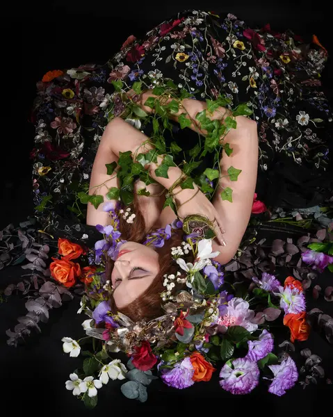 Celtic Bahar Tanrıçası Olarak Kadın Model Stüdyo Portreleri, Beltane Pagan Mitolojisinden esinlenerek Çiçek Fantezi Pelerini Kostümü. Klasik oturma pozu çiçek yatağında, izole edilmiş Karanlık Arkaplan