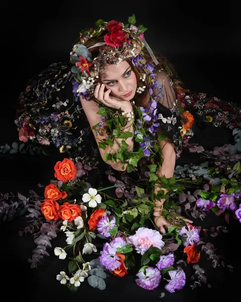 Celtic Bahar Tanrıçası Olarak Kadın Model Stüdyo Portreleri, Beltane Pagan Mitolojisinden esinlenerek Çiçek Fantezi Pelerini Kostümü. Klasik oturma pozu çiçek yatağında, izole edilmiş Karanlık Arkaplan
