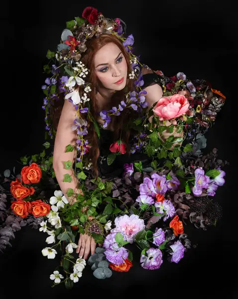 Celtic Bahar Tanrıçası Olarak Kadın Model Stüdyo Portreleri, Beltane Pagan Mitolojisinden esinlenerek Çiçek Fantezi Pelerini Kostümü. Klasik oturma pozu çiçek yatağında, izole edilmiş Karanlık Arkaplan