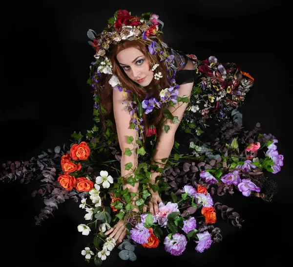 Celtic Bahar Tanrıçası Olarak Kadın Model Stüdyo Portreleri, Beltane Pagan Mitolojisinden esinlenerek Çiçek Fantezi Pelerini Kostümü. Klasik oturma pozu çiçek yatağında, izole edilmiş Karanlık Arkaplan