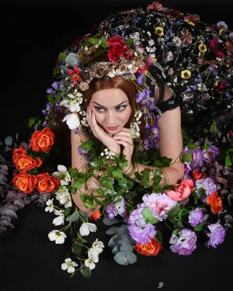 Celtic Bahar Tanrıçası Olarak Kadın Model Stüdyo Portreleri, Beltane Pagan Mitolojisinden esinlenerek Çiçek Fantezi Pelerini Kostümü. Klasik oturma pozu çiçek yatağında, izole edilmiş Karanlık Arkaplan