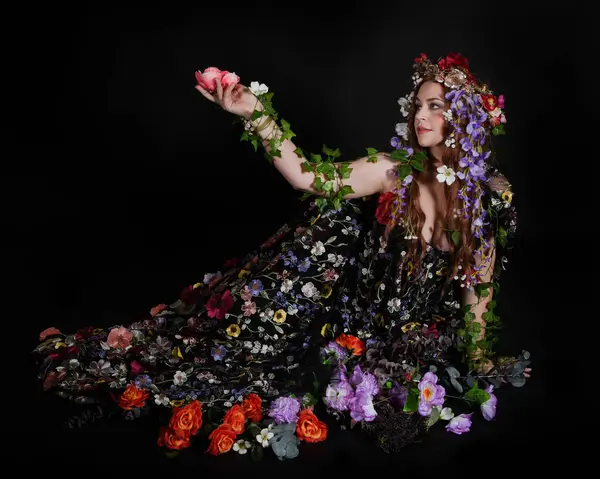 Celtic Bahar Tanrıçası Olarak Kadın Model Stüdyo Portreleri, Beltane Pagan Mitolojisinden esinlenerek Çiçek Fantezi Pelerini Kostümü. Klasik oturma pozisyonu, kollar uzanıyor. izole edilmiş Karanlık Arkaplan