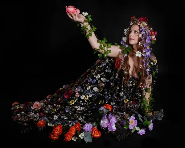 Celtic Bahar Tanrıçası Olarak Kadın Model Stüdyo Portreleri, Beltane Pagan Mitolojisinden esinlenerek Çiçek Fantezi Pelerini Kostümü. Klasik oturma pozisyonu, kollar uzanıyor. izole edilmiş Karanlık Arkaplan