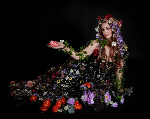 Celtic Bahar Tanrıçası Olarak Kadın Model Stüdyo Portreleri, Beltane Pagan Mitolojisinden esinlenerek Çiçek Fantezi Pelerini Kostümü. Klasik oturma pozisyonu, kollar uzanıyor. izole edilmiş Karanlık Arkaplan