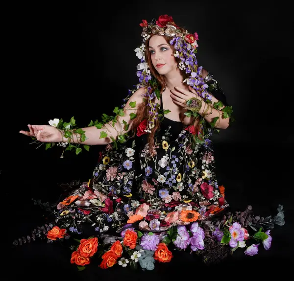 Celtic Bahar Tanrıçası Olarak Kadın Model Stüdyo Portreleri, Beltane Pagan Mitolojisinden esinlenerek Çiçek Fantezi Pelerini Kostümü. Klasik oturma pozisyonu, kollar uzanıyor. izole edilmiş Karanlık Arkaplan