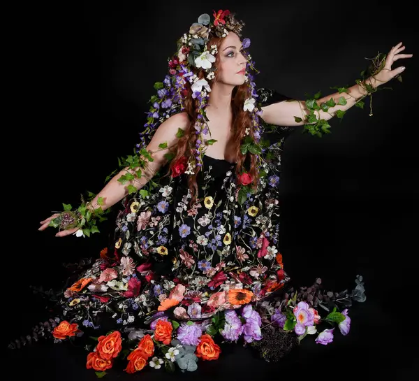 Celtic Bahar Tanrıçası Olarak Kadın Model Stüdyo Portreleri, Beltane Pagan Mitolojisinden esinlenerek Çiçek Fantezi Pelerini Kostümü. Klasik oturma pozisyonu, kollar uzanıyor. izole edilmiş Karanlık Arkaplan
