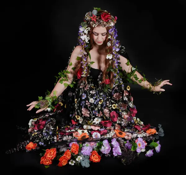 Celtic Bahar Tanrıçası Olarak Kadın Model Stüdyo Portreleri, Beltane Pagan Mitolojisinden esinlenerek Çiçek Fantezi Pelerini Kostümü. Klasik oturma pozisyonu, kollar uzanıyor. izole edilmiş Karanlık Arkaplan