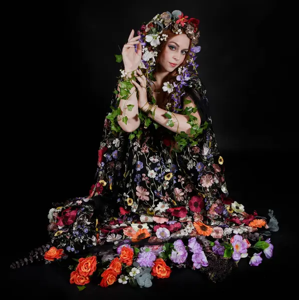 Celtic Bahar Tanrıçası Olarak Kadın Model Stüdyo Portreleri, Beltane Pagan Mitolojisinden esinlenerek Çiçek Fantezi Pelerini Kostümü. Klasik oturma pozisyonu, kollar uzanıyor. izole edilmiş Karanlık Arkaplan