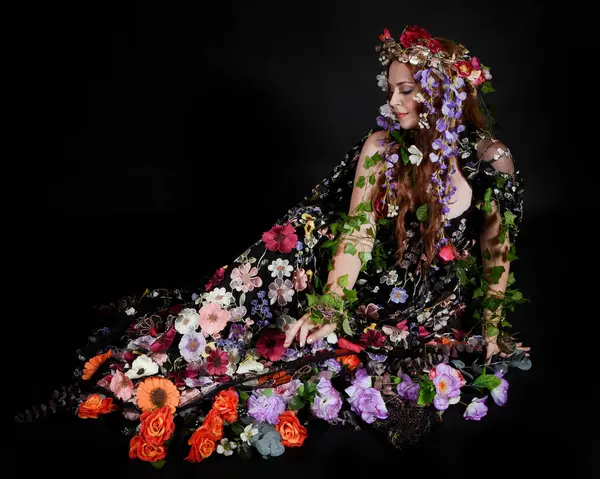 Celtic Bahar Tanrıçası Olarak Kadın Model Stüdyo Portreleri, Beltane Pagan Mitolojisinden esinlenerek Çiçek Fantezi Pelerini Kostümü. Klasik oturma pozisyonu, kollar uzanıyor. izole edilmiş Karanlık Arkaplan