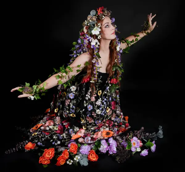 Celtic Bahar Tanrıçası Olarak Kadın Model Stüdyo Portreleri, Beltane Pagan Mitolojisinden esinlenerek Çiçek Fantezi Pelerini Kostümü. Klasik oturma pozisyonu, kollar uzanıyor. izole edilmiş Karanlık Arkaplan