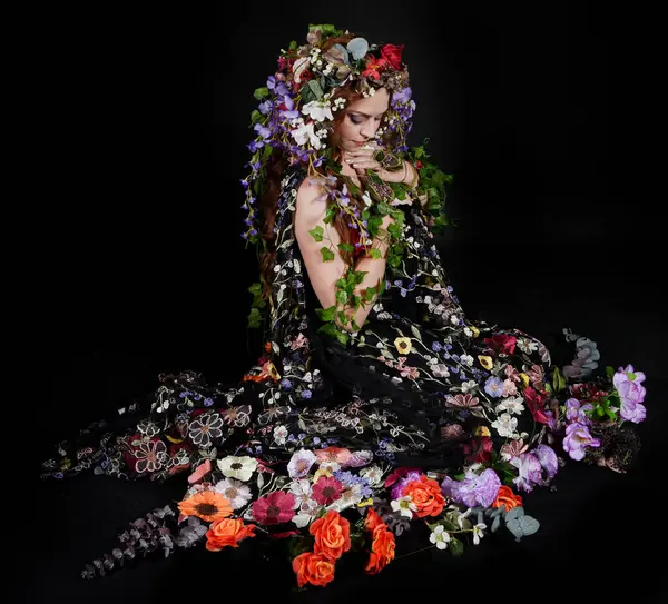 Celtic Bahar Tanrıçası Olarak Kadın Model Stüdyo Portreleri, Beltane Pagan Mitolojisinden esinlenerek Çiçek Fantezi Pelerini Kostümü. Klasik oturma pozisyonu, kollar uzanıyor. izole edilmiş Karanlık Arkaplan