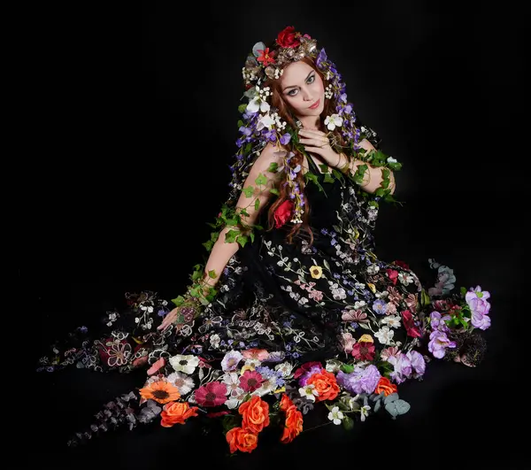 Celtic Bahar Tanrıçası Olarak Kadın Model Stüdyo Portreleri, Beltane Pagan Mitolojisinden esinlenerek Çiçek Fantezi Pelerini Kostümü. Klasik oturma pozisyonu, kollar uzanıyor. izole edilmiş Karanlık Arkaplan